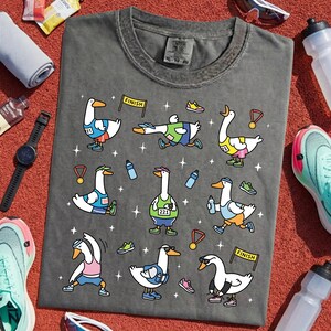 May include: Dark gray t-shirt featuring cartoon geese in athletic gear, running, stretching, and celebrating. The shirt includes the text "CRIMINAL WEAR" and size information. Accessories like shoes, water bottles, and a watch are scattered around the shirt.