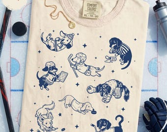Könnte beinhalten: Ein elfenbeinfarbenes T-Shirt mit einem marineblauen Grafikdruck verschiedener Dackel, die Eishockey spielen. Die Hunde tragen Eishockeyausrüstung wie Helme, Trikots und Schlittschuhe, ergänzt durch verstreute Sternmotive auf dem Stoff.
