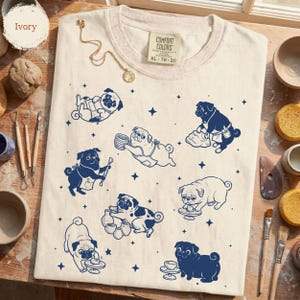 May include: An ivory t-shirt featuring a navy blue graphic print of multiple pugs engaged in pottery activities, including sculpting and using a pottery wheel, interspersed with small star shapes. The shirt has a Comfort Colors brand label.