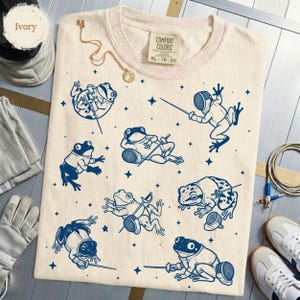 May include: An ivory t-shirt featuring a repeating pattern of dark blue illustrations of frogs engaged in fencing. The frogs are depicted in various poses, wearing fencing masks and holding foils, interspersed with small star shapes. The shirt has a tag.