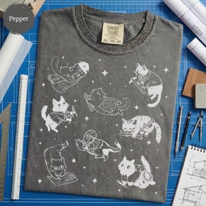 May include: Pepper-colored Comfort Colors t-shirt with white illustrations of cats in various architect and construction poses. The shirt has a crew neck and a tag that reads XL 78-20.