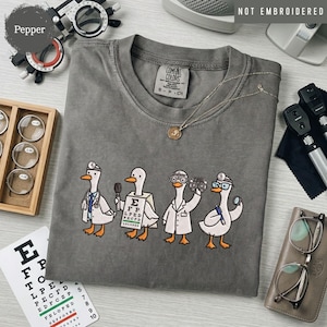 May include: A charcoal gray t-shirt featuring a graphic of four white ducks dressed as eye doctors. The ducks are wearing medical gear, holding an eye chart, and using optometry tools. The shirt has a crew neck and a Comfort Colors brand label.