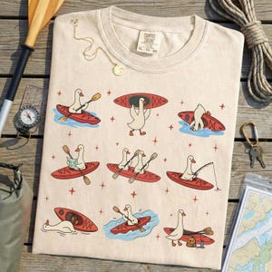 May include: Cream-colored t-shirt featuring a whimsical design of cartoon geese kayaking in red kayaks. The geese are depicted paddling, fishing, and carrying kayaks. The shirt has the text "COMFORT COLORS XL 70-20".