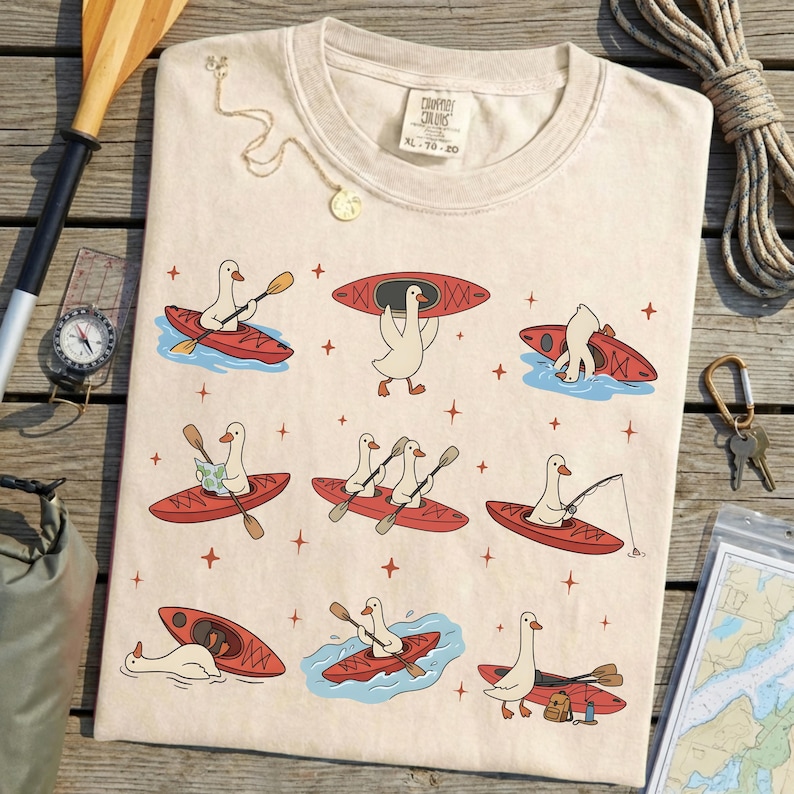 May include: Cream-colored t-shirt featuring a whimsical design of cartoon geese kayaking in red kayaks. The geese are depicted paddling, fishing, and carrying kayaks. The shirt has the text "COMFORT COLORS XL 70-20".