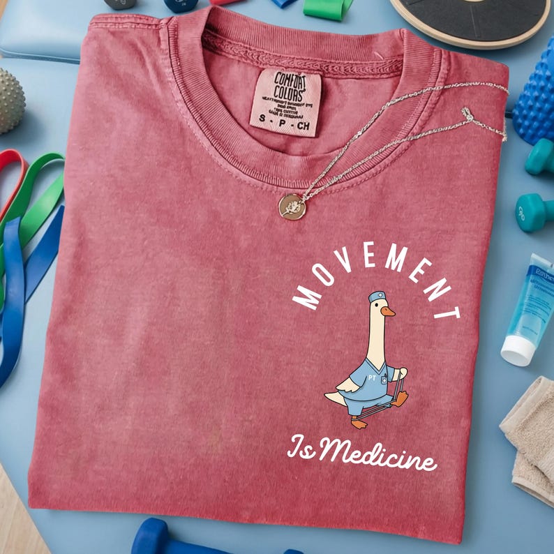 May include: A red Comfort Colors t-shirt with the words "MOVEMENT Is Medicine" and a cartoon goose wearing a doctor's uniform. A silver necklace with a pendant is on the shirt. Exercise equipment is in the background.
