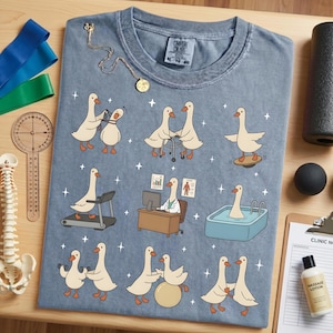 Pode incluir: Uma camiseta Comfort Colors azul clara com gansos de desenho animado em v&aacute;rios cen&aacute;rios de fisioterapia. Os gansos s&atilde;o retratados usando uma esteira, equilibrando-se e usando pesos. A camisa tamb&eacute;m tem uma etiqueta pequena.
