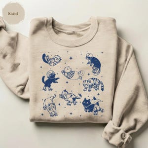 May include: Sand-coloured sweatshirt with a navy blue graphic of cats on roller skates. The cats are shown in different poses, wearing helmets and with star accents. The sweatshirt has a crew neck and long sleeves.