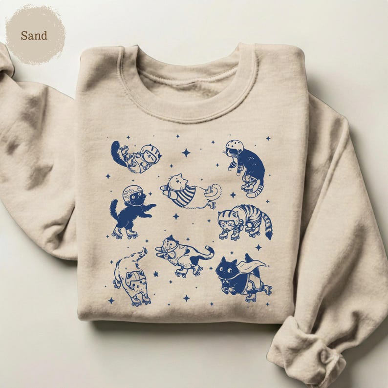 May include: Sand-coloured sweatshirt with a navy blue graphic of cats on roller skates. The cats are shown in different poses, wearing helmets and with star accents. The sweatshirt has a crew neck and long sleeves.