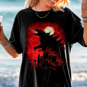 May include: Black t-shirt featuring a Godzilla graphic. The design shows the silhouette of Godzilla against a red background with a full moon and city skyline. The shirt is a comfortable, casual wear item.