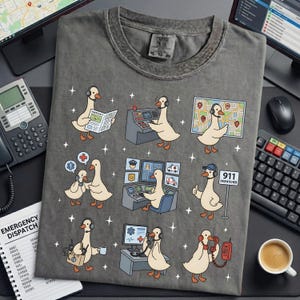 May include: Gray t-shirt featuring cartoon geese in various emergency dispatcher roles, including reading a newspaper, using a computer, and answering a phone. The shirt has the text "911 DISPATCHER".