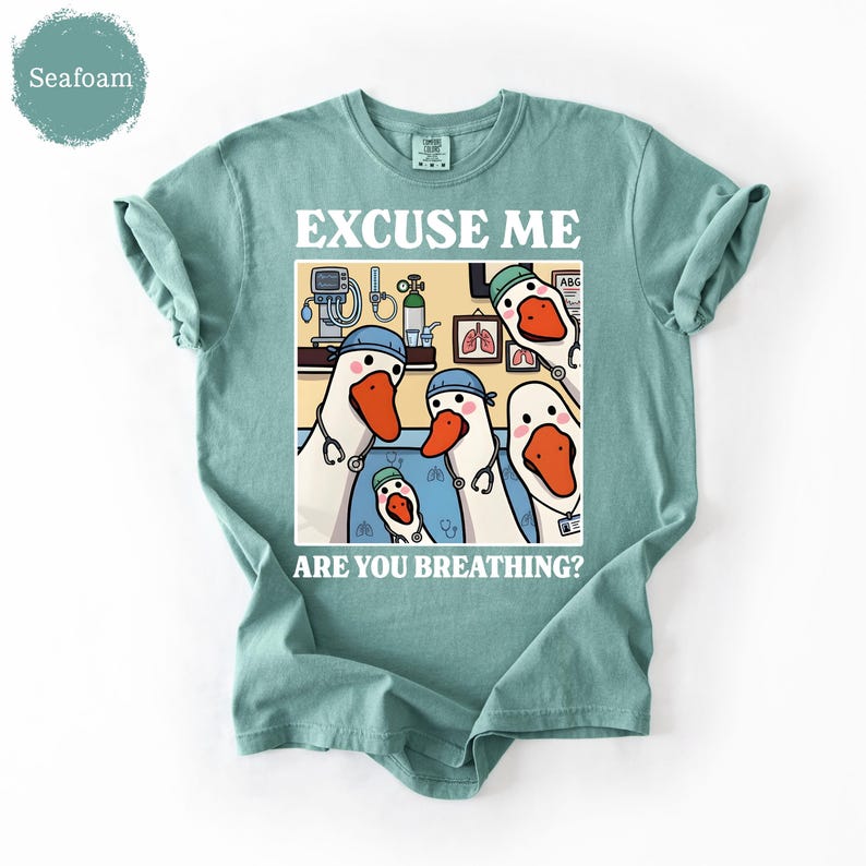 May include: Seafoam green t-shirt with a humorous graphic. The graphic features cartoon geese dressed as medical professionals, with the text "EXCUSE ME ARE YOU BREATHING?" in white. The geese have stethoscopes and are in a medical setting.