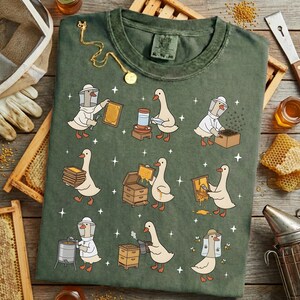 Puede incluir: Camiseta verde con una divertida ilustraci&oacute;n de gansos vestidos de apicultores. El dise&ntilde;o muestra gansos trabajando con panales, colmenas y herramientas de apicultura. Un collar dorado con un colgante est&aacute; sobre la camiseta.