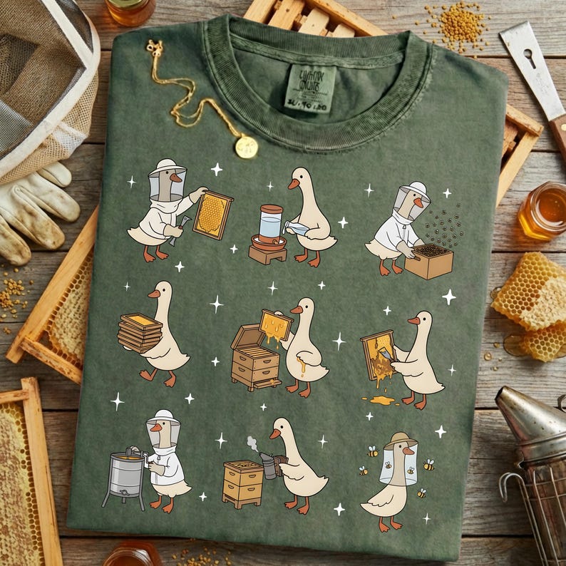 Puede incluir: Camiseta verde con una divertida ilustraci&oacute;n de gansos vestidos de apicultores. El dise&ntilde;o muestra gansos trabajando con panales, colmenas y herramientas de apicultura. Un collar dorado con un colgante est&aacute; sobre la camiseta.