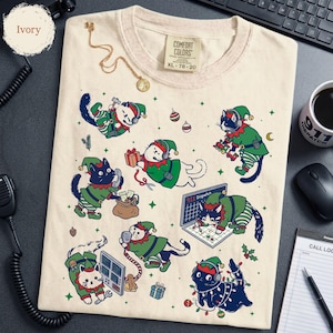 May include: An ivory t-shirt featuring a graphic print of multiple cats dressed as Christmas elves. The cats are depicted performing various dispatch tasks, including using headsets, radios, and computers with 911 dispatch screens visible.