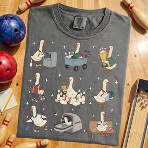 May include: Dark gray t-shirt featuring cartoon geese playing bowling. The geese are depicted in various bowling scenarios, including holding trophies, bowling, and sitting on a bench. The shirt has a vintage, worn look.