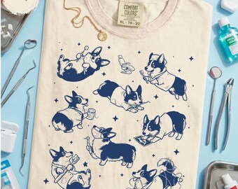 Könnte beinhalten: Ein elfenbeinfarbenes T-Shirt mit einem dunkelblauen Grafikdruck verschiedener Corgi-Hunde, die als Zahnärzte und Dentalhygieniker gekleidet sind und Aufgaben wie Zähneputzen ausführen, umgeben von kleinen Sternsymbolen und einem Comfort Colors-Etikett.