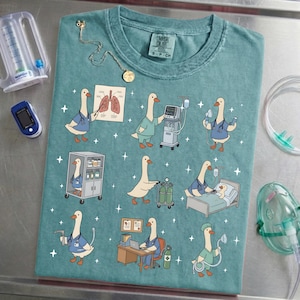 May include: Teal-colored t-shirt featuring cartoon geese in medical attire, performing various healthcare tasks. The geese are depicted with stethoscopes, oxygen tanks, and medical equipment. A necklace and medical devices are also visible.