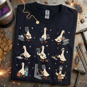 May include: Black t-shirt featuring a repeating pattern of cartoon geese welding. Each goose is depicted with welding tools and equipment. A gold necklace with a pendant is draped on the shirt.