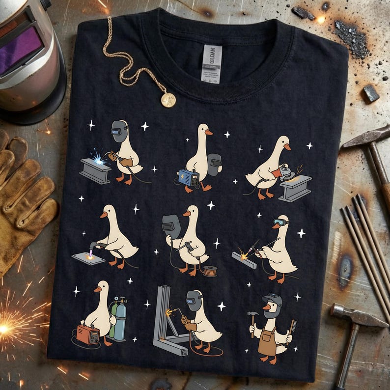 May include: Black t-shirt featuring a repeating pattern of cartoon geese welding. Each goose is depicted with welding tools and equipment. A gold necklace with a pendant is draped on the shirt.