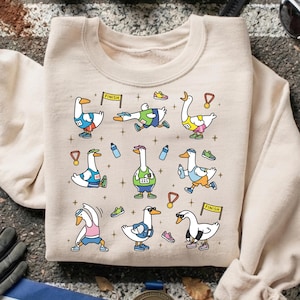 May include: Beige sweatshirt featuring a whimsical design of cartoon geese participating in a race. The geese are depicted in various poses, wearing running gear, and holding medals. The design includes water bottles, running shoes, and finish line banners.