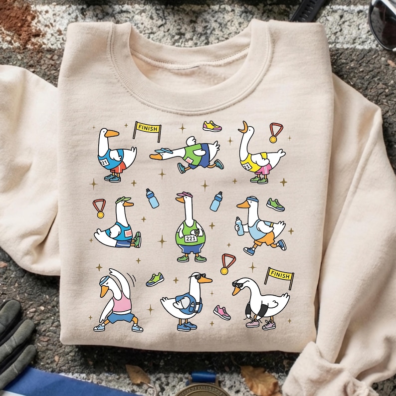 May include: Beige sweatshirt featuring a whimsical design of cartoon geese participating in a race. The geese are depicted in various poses, wearing running gear, and holding medals. The design includes water bottles, running shoes, and finish line banners.