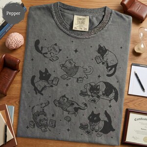 May include: A charcoal gray crew neck t-shirt featuring a black graphic print of multiple cats in various poses, some wearing glasses or lab coats, surrounded by small star shapes and books. The shirt has a Comfort Colors brand label at the neck.