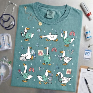 May include: Teal Comfort Colors t-shirt featuring cartoon geese in medical scrubs, using stethoscopes, oxygen masks, and other medical equipment. The shirt has a crew neck and a small tag at the top.