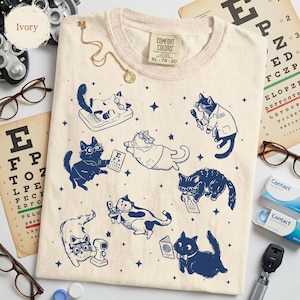 May include: Ivory-colored Comfort Colors t-shirt with a navy blue graphic of cats as optometrists. The cats are wearing glasses and examining eye charts. The shirt has a crew neck and a tag that says XL 78-20.