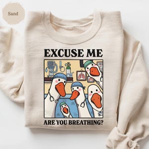 May include: Sand-colored sweatshirt featuring a humorous graphic of cartoon geese dressed as medical professionals. The geese are wearing scrubs and stethoscopes, with the text "EXCUSE ME ARE YOU BREATHING?" above and below the illustration.