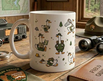 Könnte beinhalten: Ein weißer Keramikbecher mit einem wiederkehrenden Muster aus Zeichentrickenten, die als Parkranger gekleidet sind. Die Enten sind beim Wandern, mit Rucksäcken, Karten, Schmetterlingsnetzen und an Lagerfeuern dargestellt, ergänzt durch kleine Bäume und Sterne.