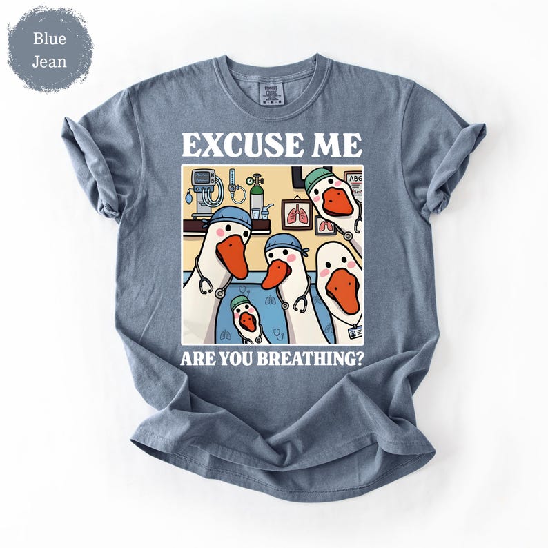 May include: Blue jean colored t-shirt with a humorous graphic. The graphic features cartoon geese dressed as medical professionals, with the text "EXCUSE ME ARE YOU BREATHING?"