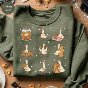 May include: Military green sweatshirt featuring a whimsical design of geese and waffles. The graphic includes geese with waffles, in a jar, and on a picnic blanket. The sweatshirt has a crew neck and long sleeves. The text "Military Green" is in the upper left corner.