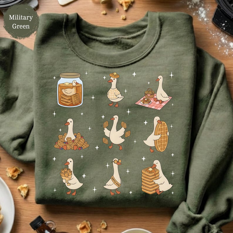May include: Military green sweatshirt featuring a whimsical design of geese and waffles. The graphic includes geese with waffles, in a jar, and on a picnic blanket. The sweatshirt has a crew neck and long sleeves. The text "Military Green" is in the upper left corner.
