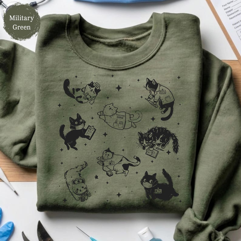 May include: Military green sweatshirt featuring a pattern of cartoon cats in various medical roles, including wearing masks and holding clipboards. The cats are black and white, and the design includes small star accents.