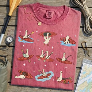 May include: A red-pink t-shirt featuring cartoon geese kayaking, fishing, and carrying kayaks. The design includes a compass, rope, and a map. The shirt has a crew neck and short sleeves. The shirt is made of a soft, comfortable material.