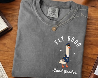 May include: A gray Comfort Colors t-shirt featuring a cartoon goose in a pilot uniform with the words "FLY GOOD" and "Land Gooder". The shirt has a crew neck and short sleeves. A silver necklace with a pendant is on the shirt.