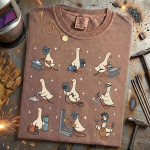 May include: Brown t-shirt featuring a repeating pattern of cartoon geese welding. Each goose is depicted with welding tools and equipment. The shirt has a crew neck and a tag that reads "Comfort Colors XL-TG 20". A gold necklace is draped on the shirt.