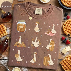 May include: Espresso-colored t-shirt with a whimsical design featuring cartoon geese interacting with waffles. The geese are depicted in various scenes, including enjoying waffles and wearing waffle-themed accessories. The shirt has a Comfort Colors tag, size XL-78-20.