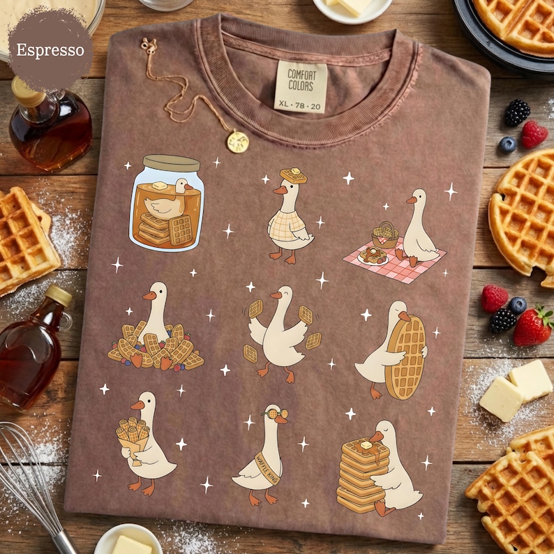 May include: Espresso-colored t-shirt with a whimsical design featuring cartoon geese interacting with waffles. The geese are depicted in various scenes, including enjoying waffles and wearing waffle-themed accessories. The shirt has a Comfort Colors tag, size XL-78-20.