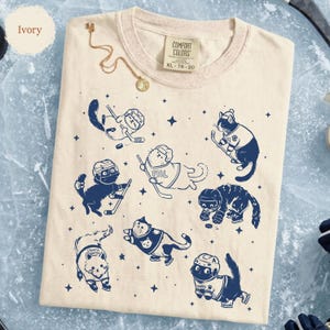May include: Ivory-colored t-shirt with a crew neck and a graphic of cats playing hockey. The cats are wearing helmets and jerseys, and the design includes hockey sticks and stars. The shirt has a Comfort Colors tag, size XL 78-20.