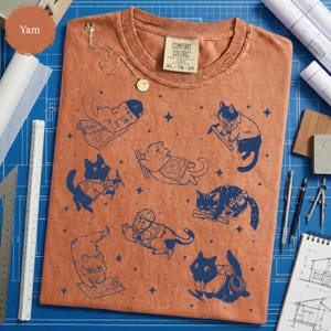May include: A yam-colored Comfort Colors XL t-shirt featuring a navy blue graphic of cats in various construction poses. The cats are depicted with tools and blueprints. A gold necklace is draped on the shirt.