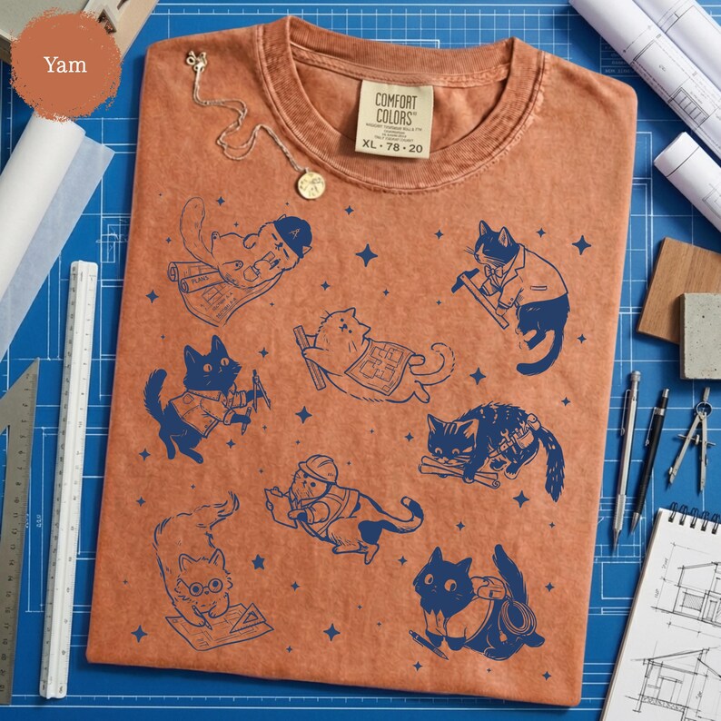 May include: A yam-colored Comfort Colors XL t-shirt featuring a navy blue graphic of cats in various construction poses. The cats are depicted with tools and blueprints. A gold necklace is draped on the shirt.