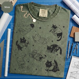 May include: Moss green Comfort Colors XL t-shirt featuring black cat illustrations in various construction-related poses. The shirt has a crew neck and short sleeves. A gold necklace is draped on the shirt.