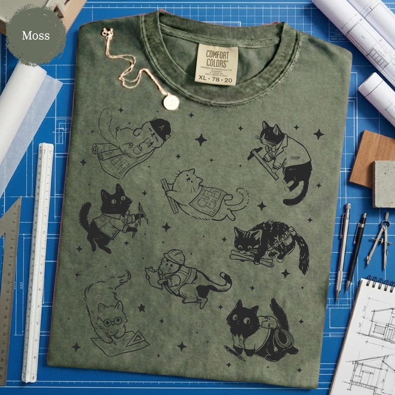 May include: Moss green Comfort Colors XL t-shirt featuring black cat illustrations in various construction-related poses. The shirt has a crew neck and short sleeves. A gold necklace is draped on the shirt.