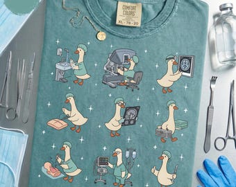 Könnte beinhalten: Seafoam-grünes Comfort Colors T-Shirt mit einer skurrilen Grafik einer Gans in einer chirurgischen Umgebung. Die Gans wird bei der Durchführung verschiedener medizinischer Verfahren dargestellt. Das Shirt hat einen Rundhalsausschnitt und kurze Ärmel. Größe XL 78-20.