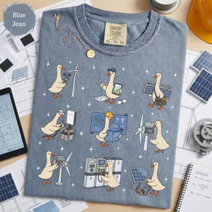 May include: Blue jean Comfort Colors t-shirt featuring a whimsical design of cartoon geese engaged in various renewable energy activities. The geese are depicted with wind turbines, solar panels, and blueprints. The shirt has a tag that reads XL 78-20.