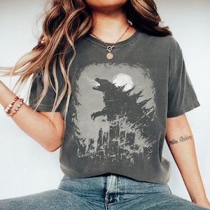 May include: Dark grey t-shirt featuring a Godzilla graphic. The design shows Godzilla roaring in front of a cityscape and a full moon. The shirt has a vintage, worn look.