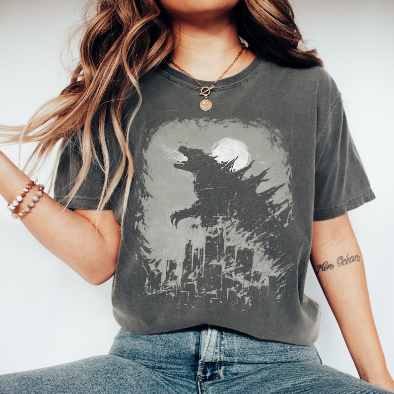 May include: Dark grey t-shirt featuring a Godzilla graphic. The design shows Godzilla roaring in front of a cityscape and a full moon. The shirt has a vintage, worn look.