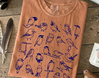May include: A terracotta-colored t-shirt featuring a collection of navy blue bird illustrations. The birds are depicted in various poses, with some perched on branches. The shirt has a classic crew neck and a tag in the upper right corner.