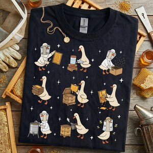 Puede incluir: Camiseta azul marino oscuro con una ilustraci&oacute;n caprichosa de gansos vestidos de apicultores. Los gansos se muestran trabajando con panales, tarros y otros equipos de apicultura. El dise&ntilde;o incluye peque&ntilde;as estrellas blancas.